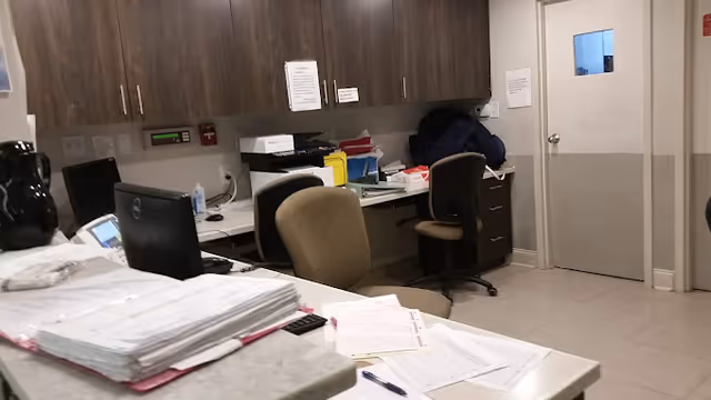 An office or administrative workspace with desks, chairs, stacks of papers, a computer monitor, and cabinets mounted on the wall. The room has a door with a small window and various office supplies scattered on the desks.