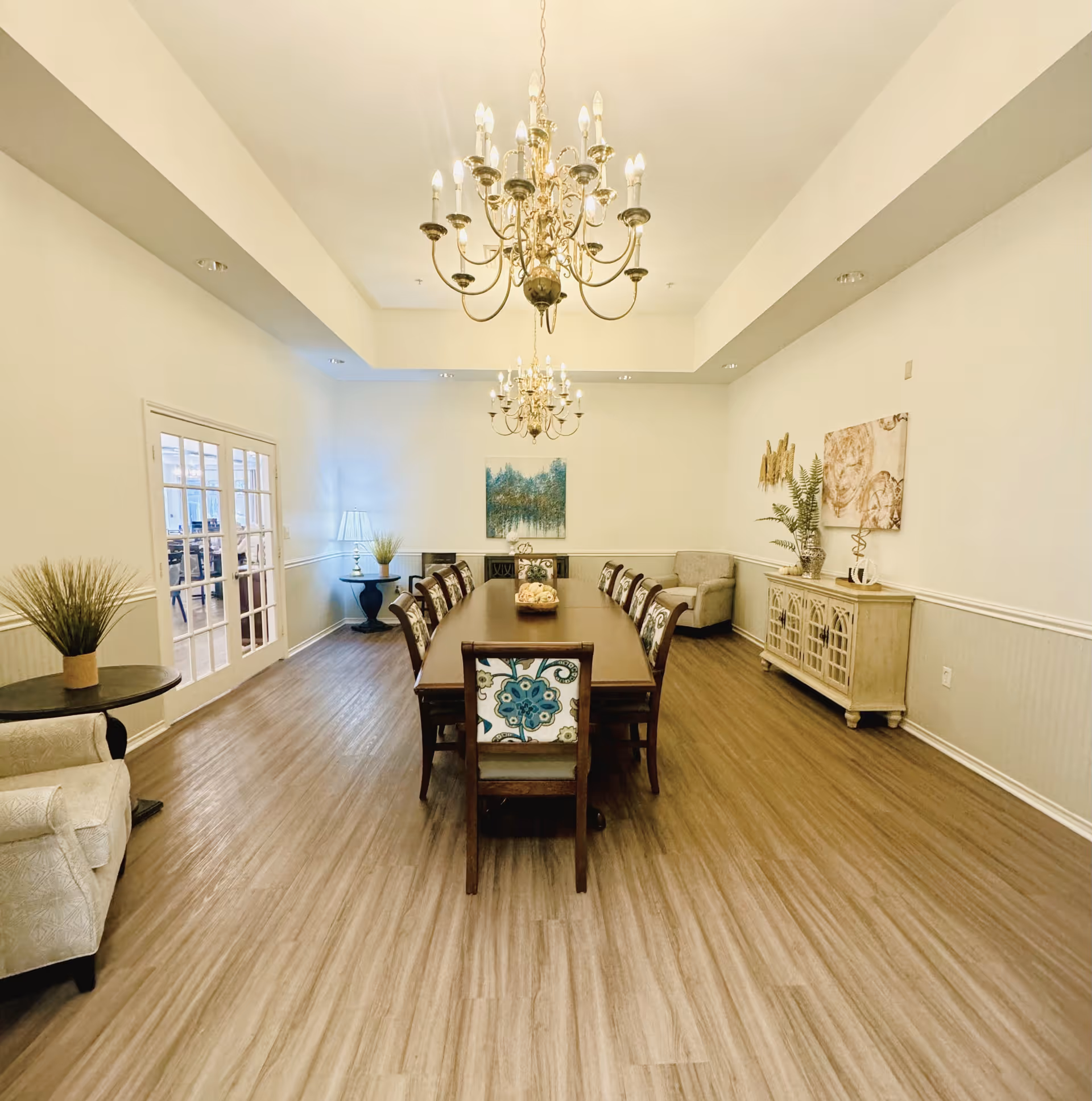 A spacious dining room with a long wooden table surrounded by chairs, two ornate chandeliers hanging from the ceiling, light-colored walls, wooden flooring, decorative plants, a sideboard with artwork above it, and a glass-paneled door on the left side.