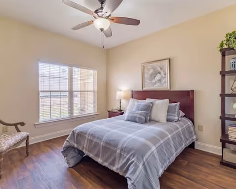A cozy bedroom with a large window letting in natural light, a bed with a gray plaid comforter and multiple pillows, a wooden nightstand with a lamp, a framed floral artwork above the bed, a wooden chair with patterned upholstery, and a tall shelving unit with decorative items and books.