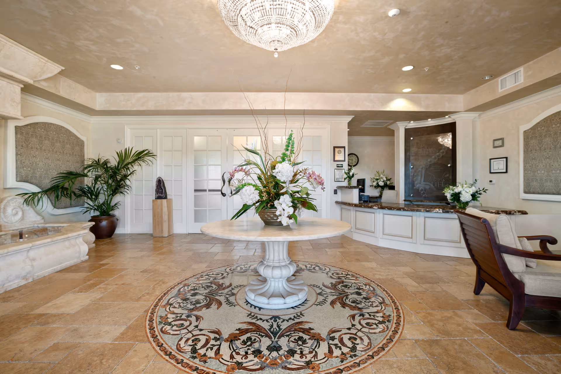 A spacious and elegant lobby area with a round marble table in the center holding a large floral arrangement. The floor features a decorative circular rug beneath the table. To the right is a reception desk with a granite countertop and a comfortable chair nearby. The walls are light-colored with framed artwork and plants adding greenery. A large chandelier hangs from the ceiling, illuminating the space.