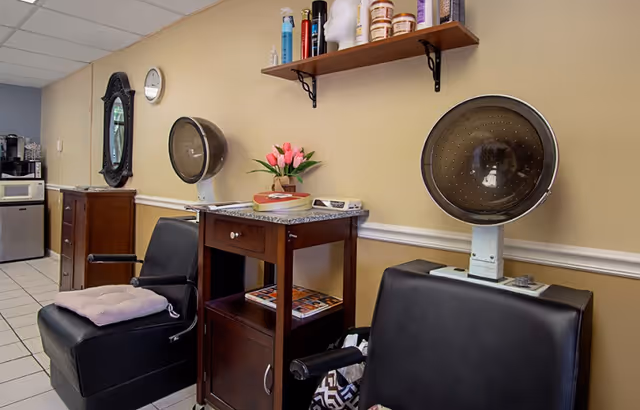Interior of a salon area with two black salon chairs, each positioned under a vintage hair dryer hood. A wooden cabinet with a granite top is placed between the chairs, holding a vase with pink tulips and some magazines on the lower shelf. Above the cabinet, a wooden shelf mounted on the wall holds various hair care products. A decorative mirror and a clock are visible on the wall in the background.