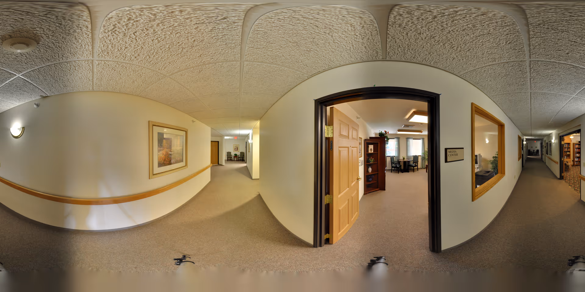 Interior hallway of a senior living facility with beige walls and carpeted floors. There is a wooden handrail along the wall and framed artwork hanging. An open door leads to a room labeled 'Media Center' with chairs, tables, and shelves inside. The hallway extends in both directions with additional doors and windows visible.
