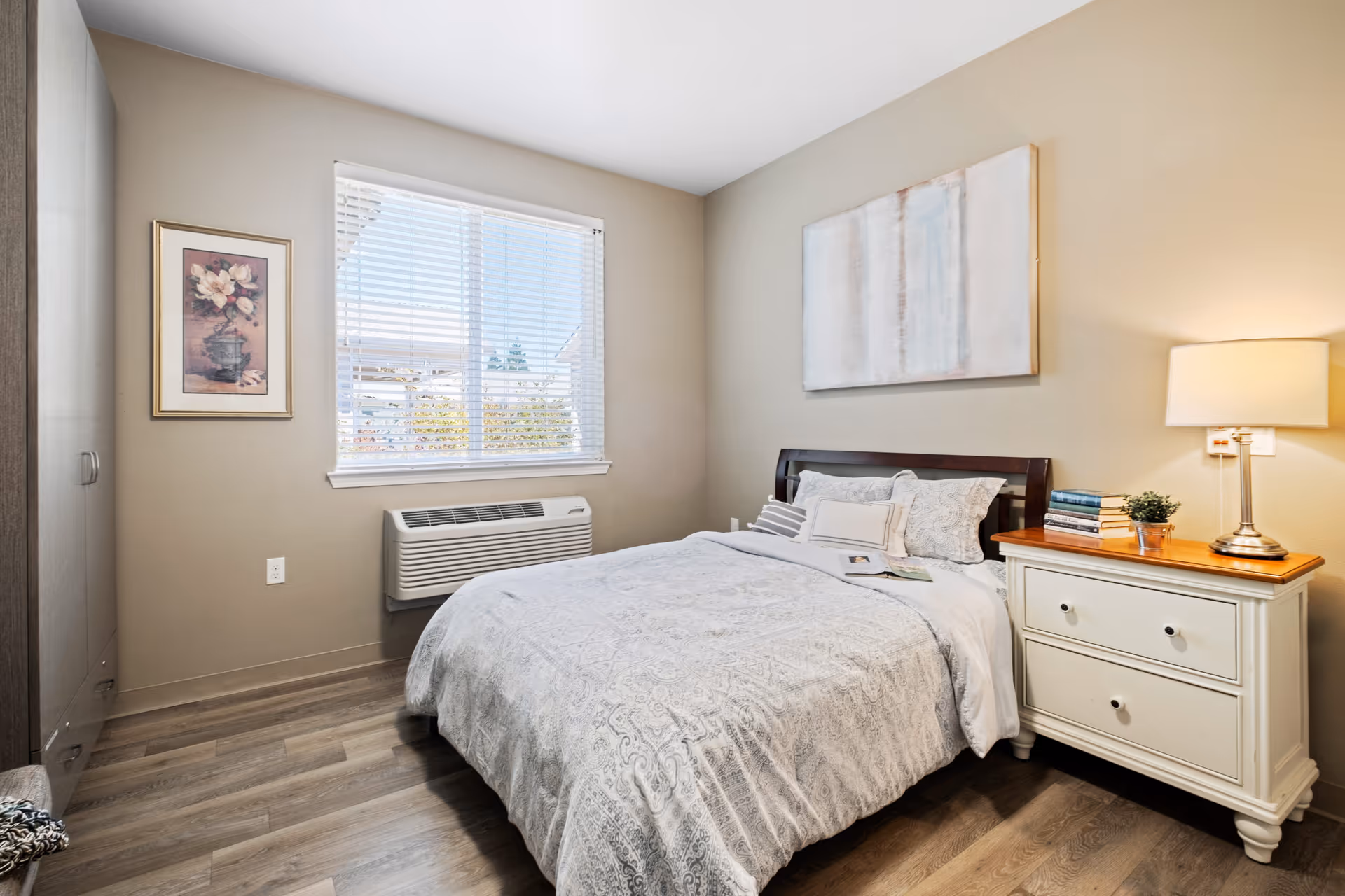 A cozy bedroom with a double bed covered in a light gray patterned comforter and multiple pillows. There is a white nightstand with two drawers next to the bed, holding a lamp, a small plant, and a stack of books. A large window with white blinds lets in natural light, and a framed floral picture hangs on the wall to the left of the window. The walls are painted beige, and the floor has wood-like vinyl planks.