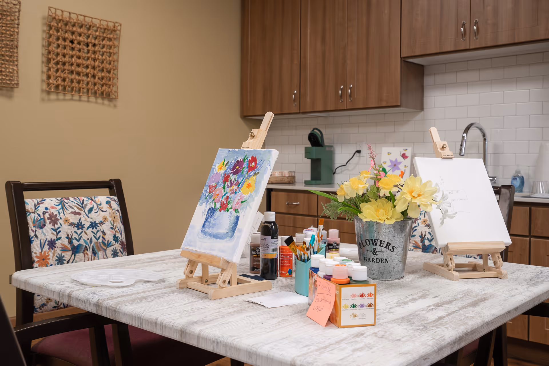 A craft table set with painting easels, paints, brushes and a vase of yellow flowers in a senior living facility's activity/kitchen area.