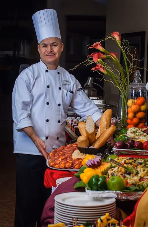 A chef in a white uniform and tall hat stands behind a buffet table laden with breads, salads, and fruit arrangements.
