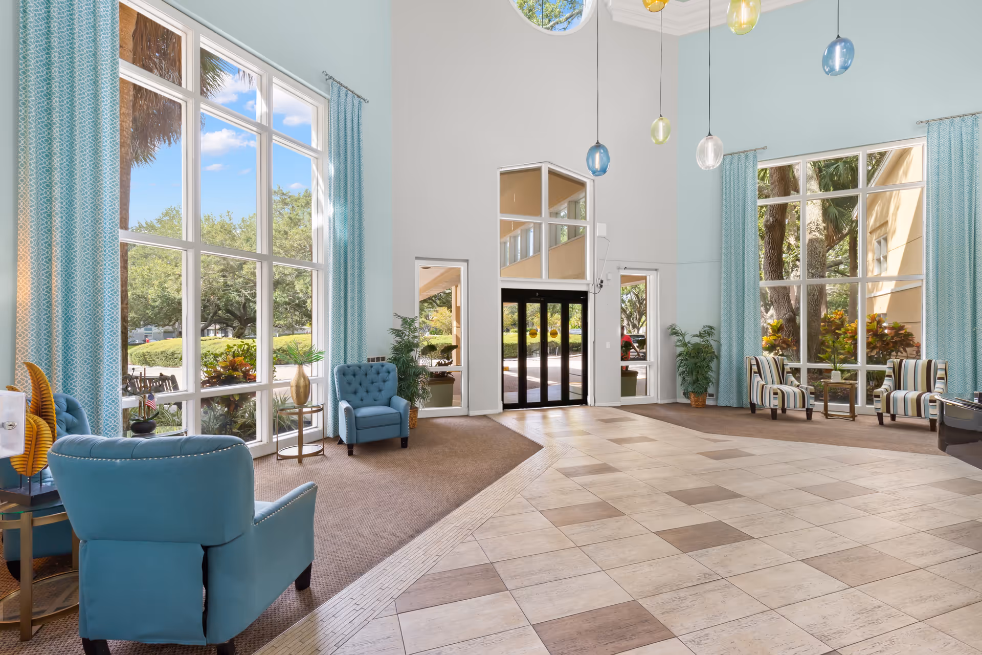 Bright and spacious lobby area with large floor-to-ceiling windows, light blue walls, and blue and striped armchairs arranged near the windows. The floor is a combination of beige and brown tiles and carpet. Several colorful pendant lights hang from the high ceiling. Outside greenery is visible through the windows.