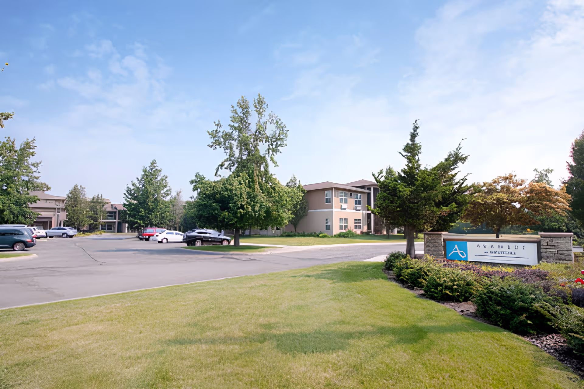 Exterior view of Wenatchee Senior Living by Cogir facility showing a parking lot with several cars, green trees, and a clear blue sky. The building is visible in the background with a sign in the foreground surrounded by landscaping.