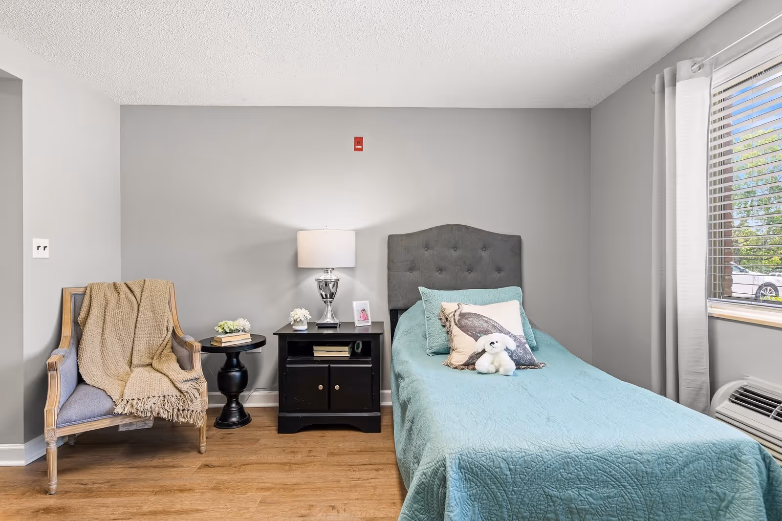 A cozy bedroom with a single bed covered in a teal quilt and decorative pillows, including one with a bird design and a small white stuffed animal. Next to the bed is a black nightstand with a silver lamp, a small flower arrangement, and a framed photo. To the left of the nightstand is a wooden armchair with a beige throw blanket draped over it. The room has light gray walls, wood flooring, and a window with white curtains and blinds letting in natural light.