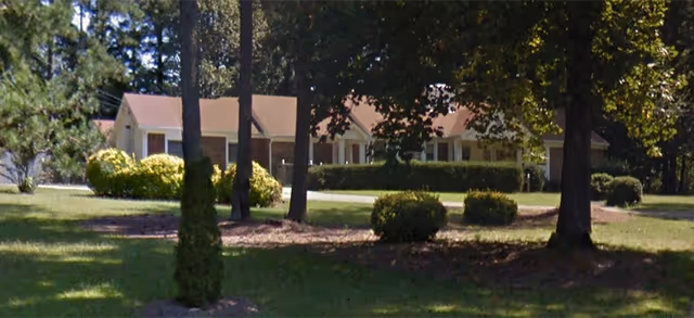 A single-story residential building with a light-colored exterior and a brown roof, surrounded by green grass, bushes, and tall trees under a clear sky.