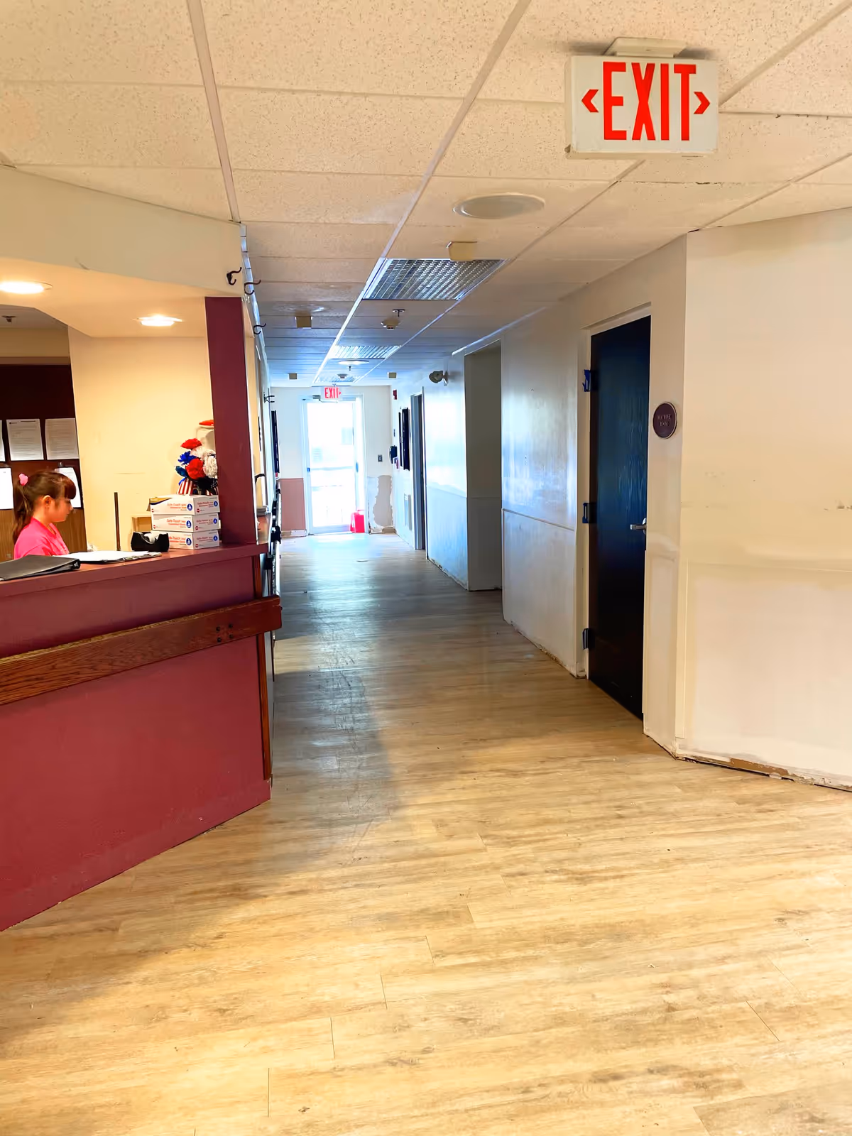 Interior hallway of a senior living facility with light wood flooring and white walls. On the left side, there is a reception desk with a person in a pink shirt standing behind it. The hallway extends toward a glass exit door with an illuminated red EXIT sign above it. There are several doors along the hallway and another EXIT sign hanging from the ceiling near the foreground.