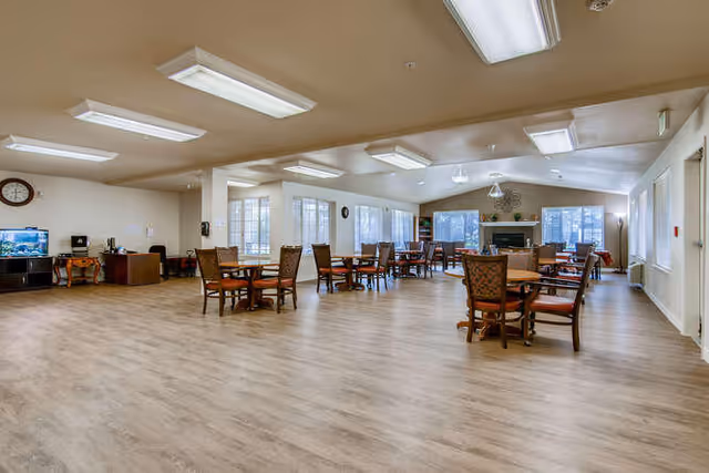 Spacious common area with multiple round wooden tables and chairs arranged on a light wood floor. Large windows allow natural light to fill the room. A fireplace is visible at the far end of the room, and there is a TV and some furniture along one wall. The ceiling has several rectangular fluorescent light fixtures.