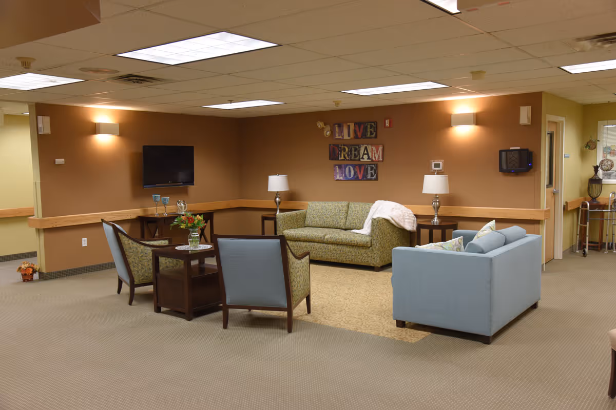 A cozy living room area in a senior living facility with two sofas, two armchairs, two side tables with lamps, a small table with a flower vase, and a wall-mounted TV. The walls are painted brown with a decorative sign that reads 'LIVE DREAM LOVE'. The room is well-lit with ceiling lights and wall sconces.