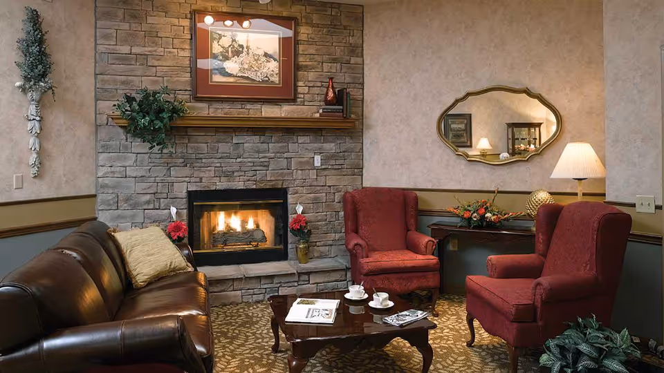 Cozy living room area with a stone fireplace, a brown leather sofa with a beige pillow, two red upholstered armchairs, a wooden coffee table with teacups and newspapers, a decorative wall mirror, a table lamp, and floral arrangements.