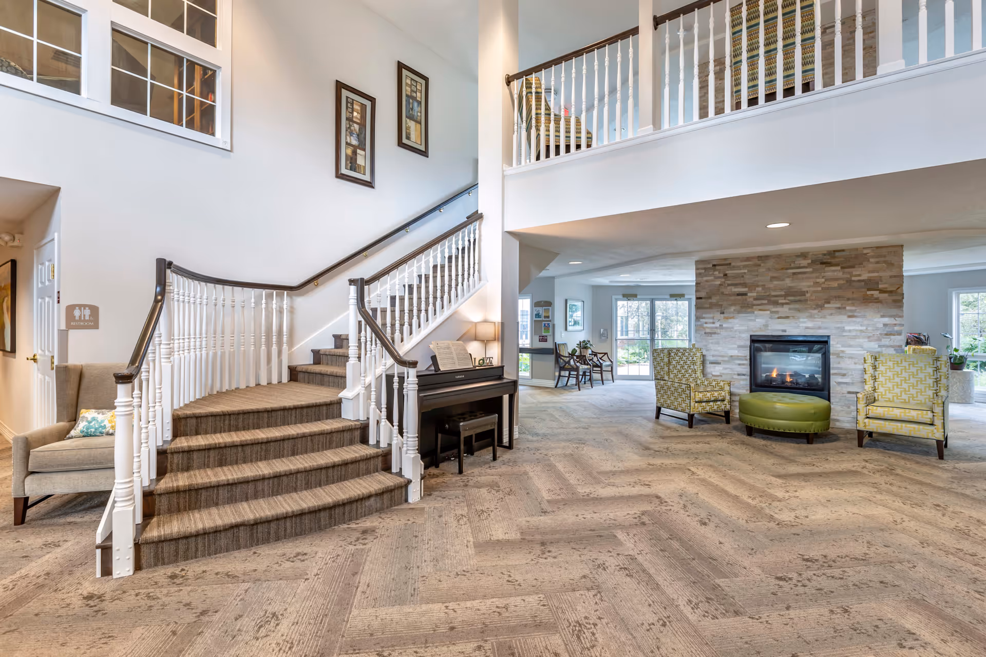 A spacious senior living facility common area featuring a carpeted staircase with white railings, a black piano with sheet music and a lamp on top, two patterned armchairs and a green ottoman in front of a stone fireplace, and additional seating near large windows letting in natural light.