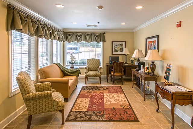 A cozy living room area with a tan sofa, two patterned armchairs, a colorful area rug, and a wooden desk with a computer and chair. The room has large windows with valance curtains letting in natural light, beige walls, and several table lamps on wooden side tables.