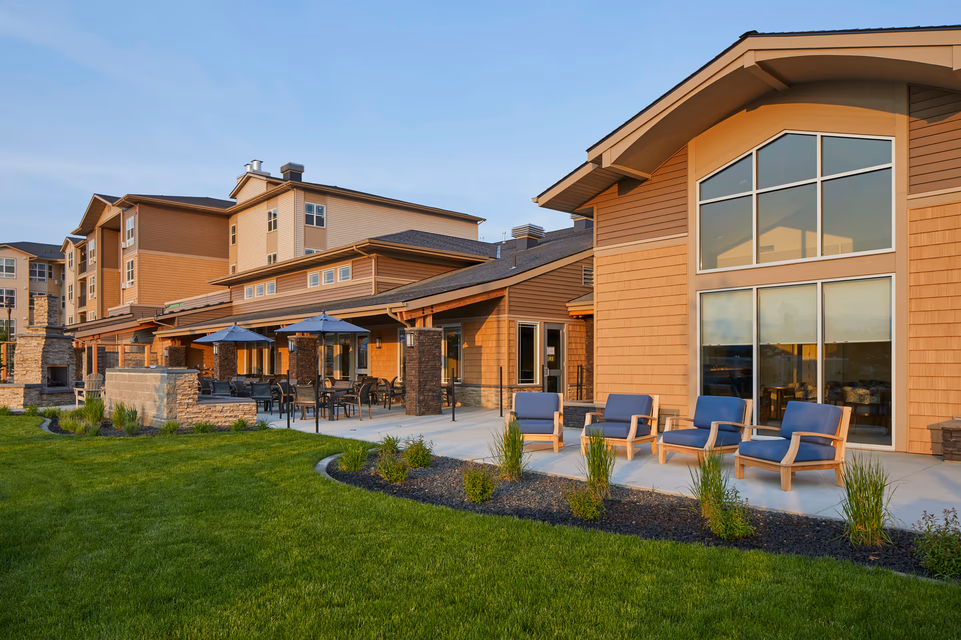 Exterior view of a multi-story senior living building with a landscaped lawn, patio seating, umbrellas, and large windows.