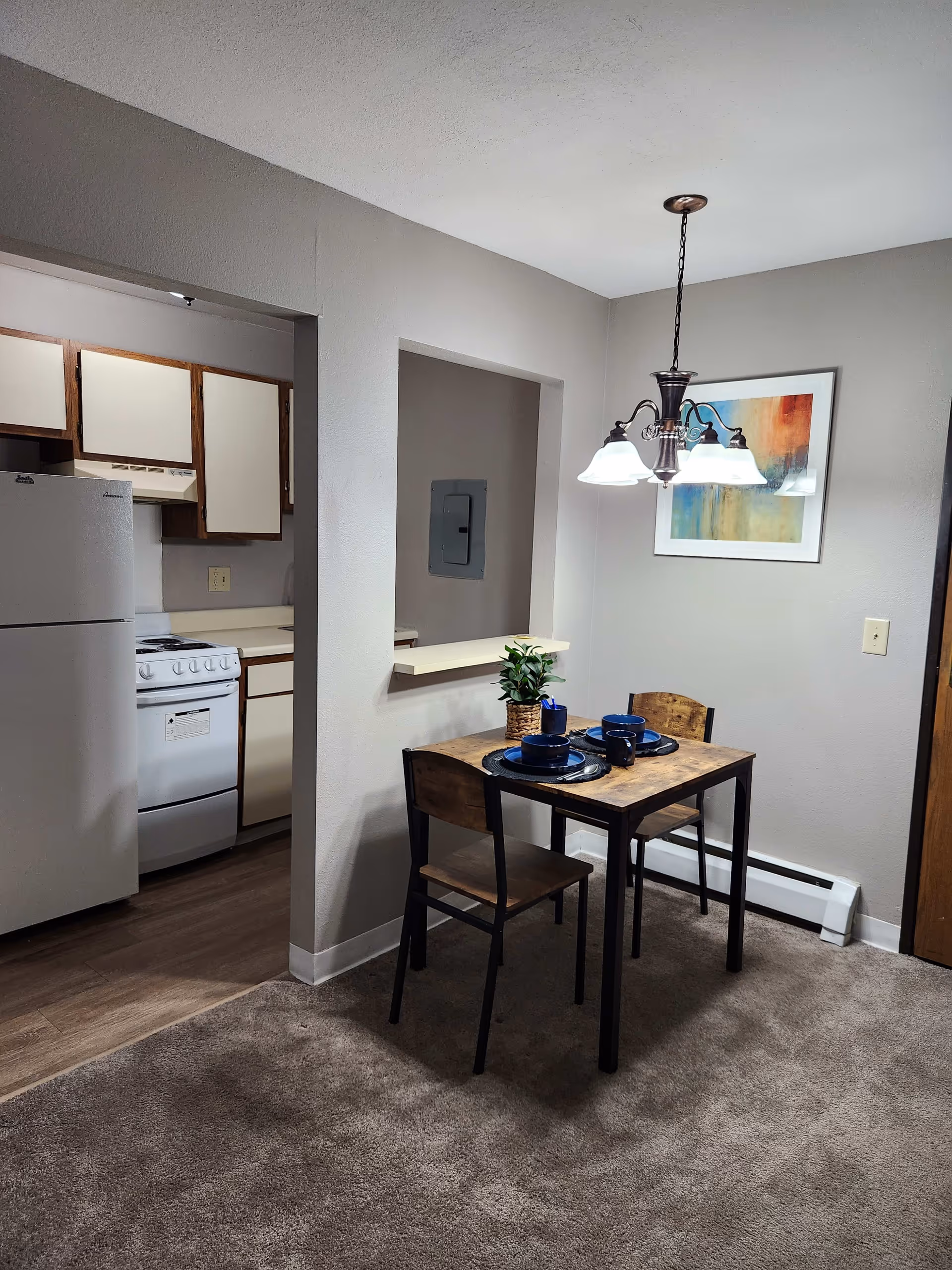 Small dining area with a wooden table set for two with blue plates and mugs, two wooden chairs, a hanging light fixture above, and a framed abstract painting on the wall. Adjacent to the dining area is a kitchen with white appliances including a refrigerator and stove, and wooden cabinets.