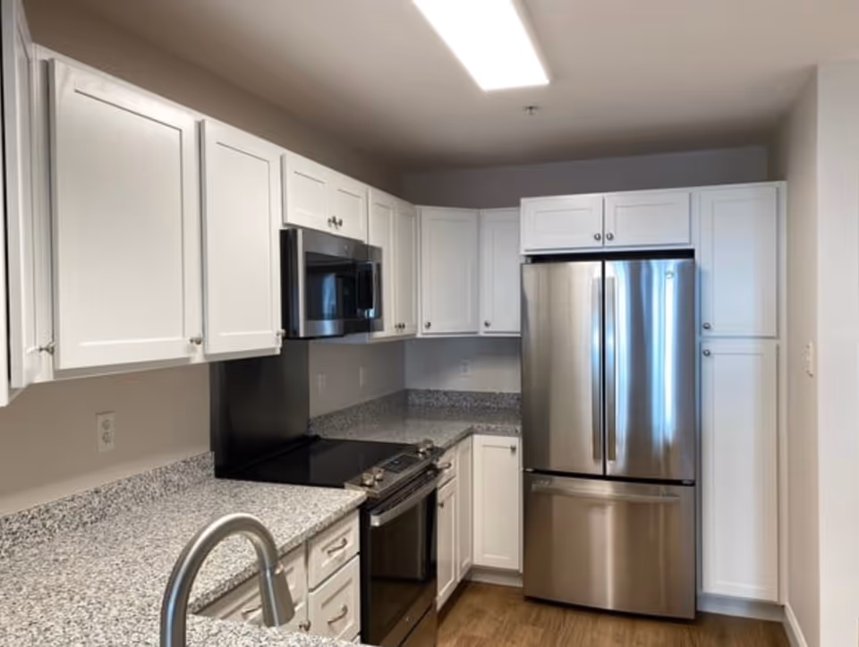 Modern kitchen with white cabinets, granite countertops, stainless steel refrigerator, stove, microwave, and a sink with a curved faucet. The floor is wooden and the ceiling has a rectangular light fixture.