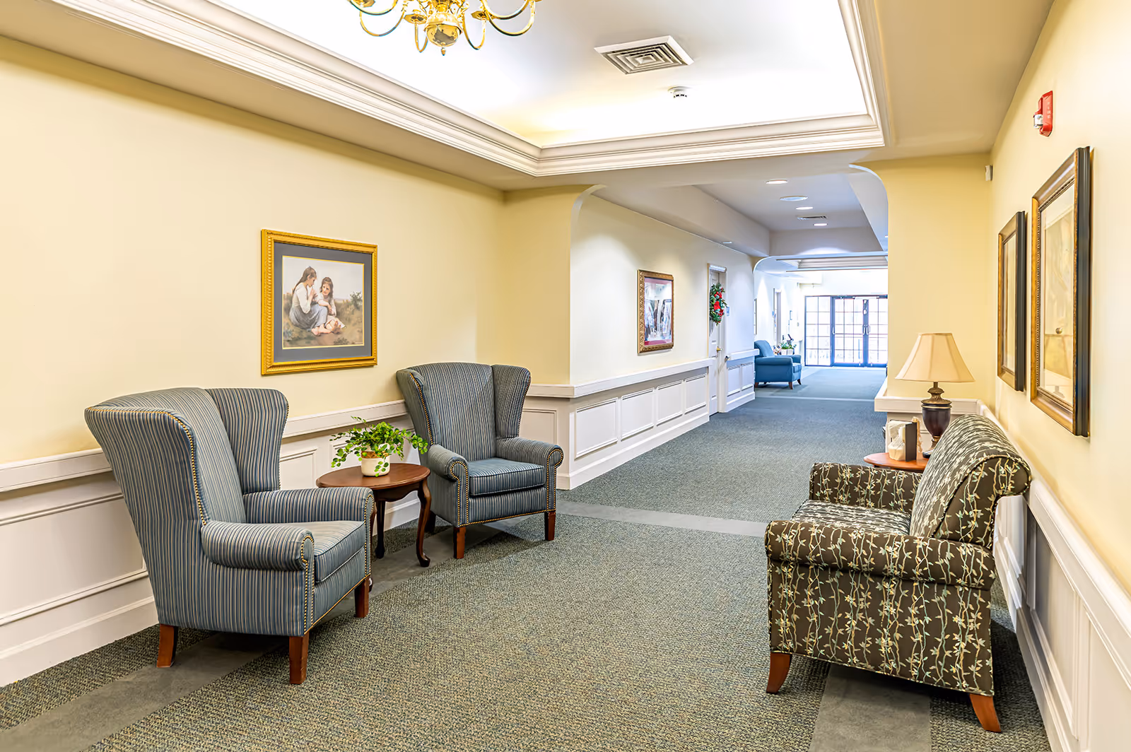 A well-lit hallway in an assisted living facility with comfortable upholstered chairs and small tables along the walls. The walls are painted light yellow with white wainscoting, and framed artwork hangs above the chairs. The carpet is greenish with a subtle pattern, and a chandelier is visible on the ceiling. The hallway extends to a glass door entrance in the background.