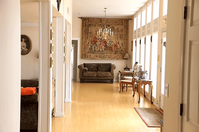 A bright hallway in a senior living facility with wooden flooring, a dark upholstered loveseat at the end, a large decorative tapestry on the wall above the loveseat, a chandelier hanging from the ceiling, and wooden tables and chairs along the right side near tall windows letting in natural light.