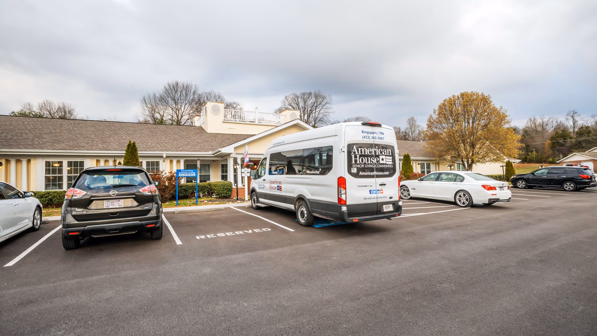 Parking lot in front of a single-story building with several parked cars and a white American House senior living community shuttle van. The building has beige siding, white trim, and a small porch entrance. Trees with autumn foliage are visible in the background under a cloudy sky.