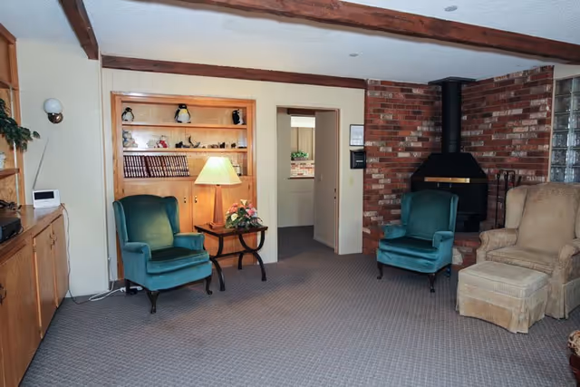 A cozy living room with two teal armchairs, a beige armchair with an ottoman, a brick fireplace with a black chimney, wooden beams on the ceiling, a wooden built-in bookshelf with decorative items, a side table with a lamp and flowers, and a doorway leading to another room.