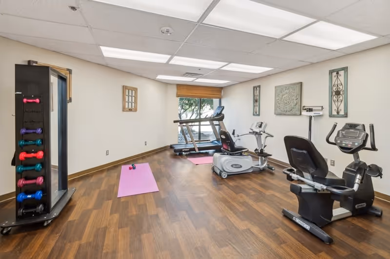 Small fitness room with treadmills, stationary bikes, dumbbells, and yoga mats on wood flooring.