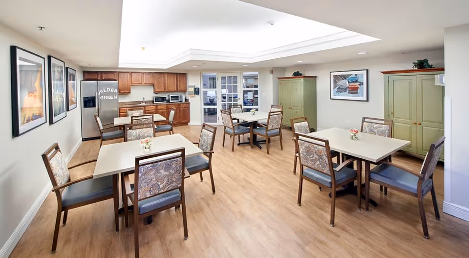 A bright dining room with four square tables, each surrounded by four chairs with patterned upholstery. The room has light wood flooring, a kitchen area with wooden cabinets, a refrigerator with the words 'Aldea Green' on it, and two green storage cabinets against the far wall. There are framed pictures on the walls and a skylight providing natural light.