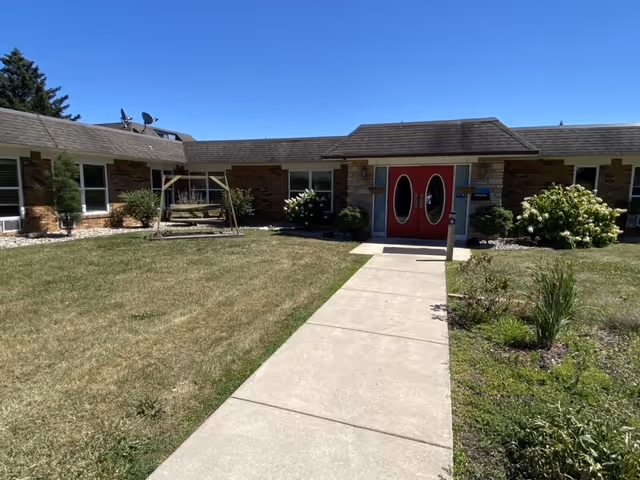 Single-story brick building courtyard with a concrete walkway leading to red double doors, a lawn, wooden swing, and landscaping under a clear blue sky.