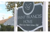 Outdoor sign for Saint Francis Home with the tagline 'Where Love Never Grows Old' displayed on a dark background with white text, surrounded by trees and a building in the background.