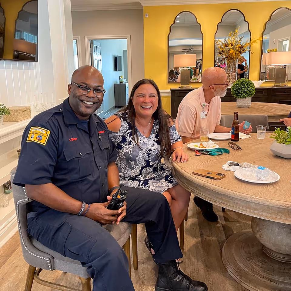 Three smiling adults sit around a round table with plates and drinks in a bright communal dining area of a senior care home.