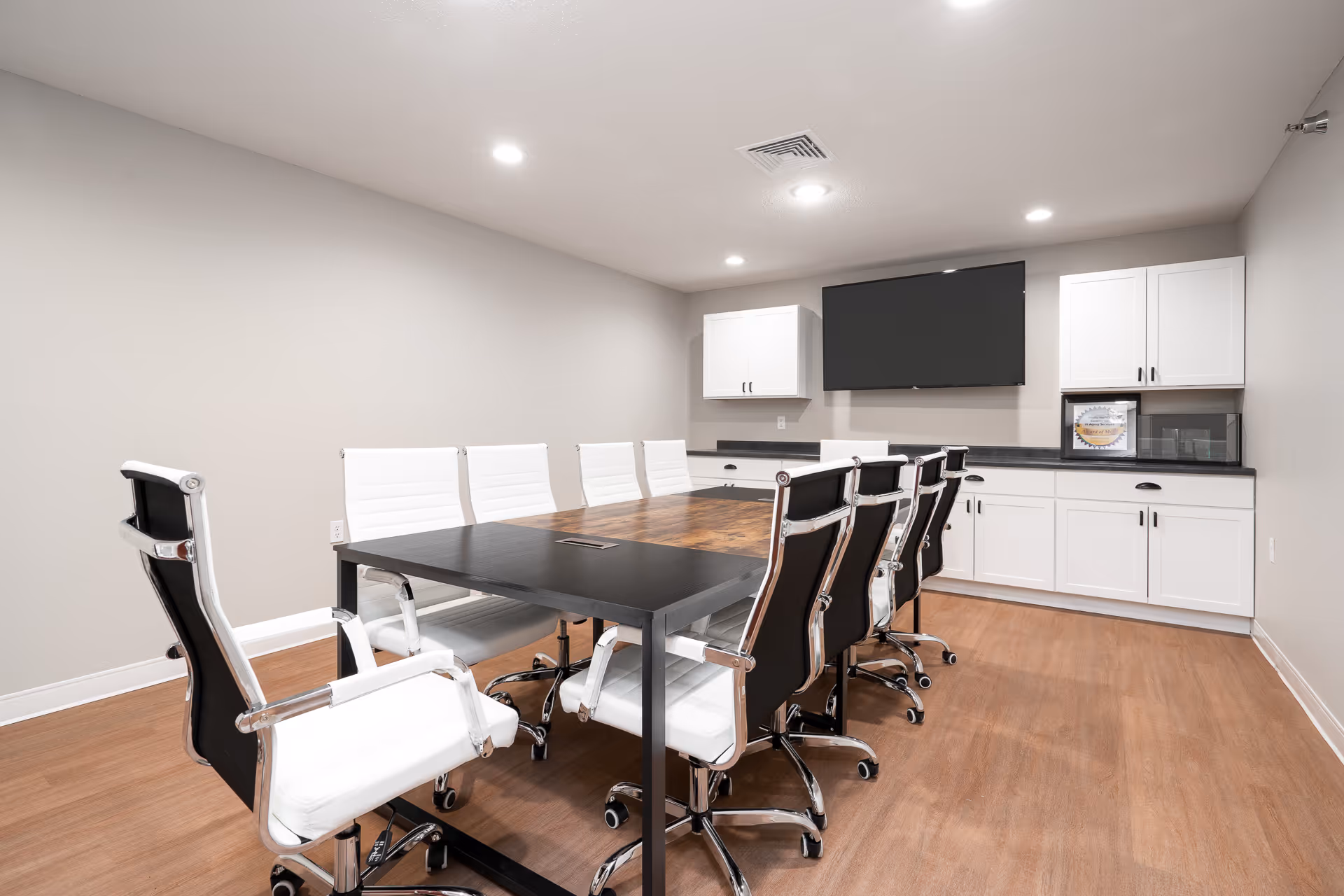 Modern conference room with a long table surrounded by white and black office chairs, a wall-mounted TV, and white cabinets.