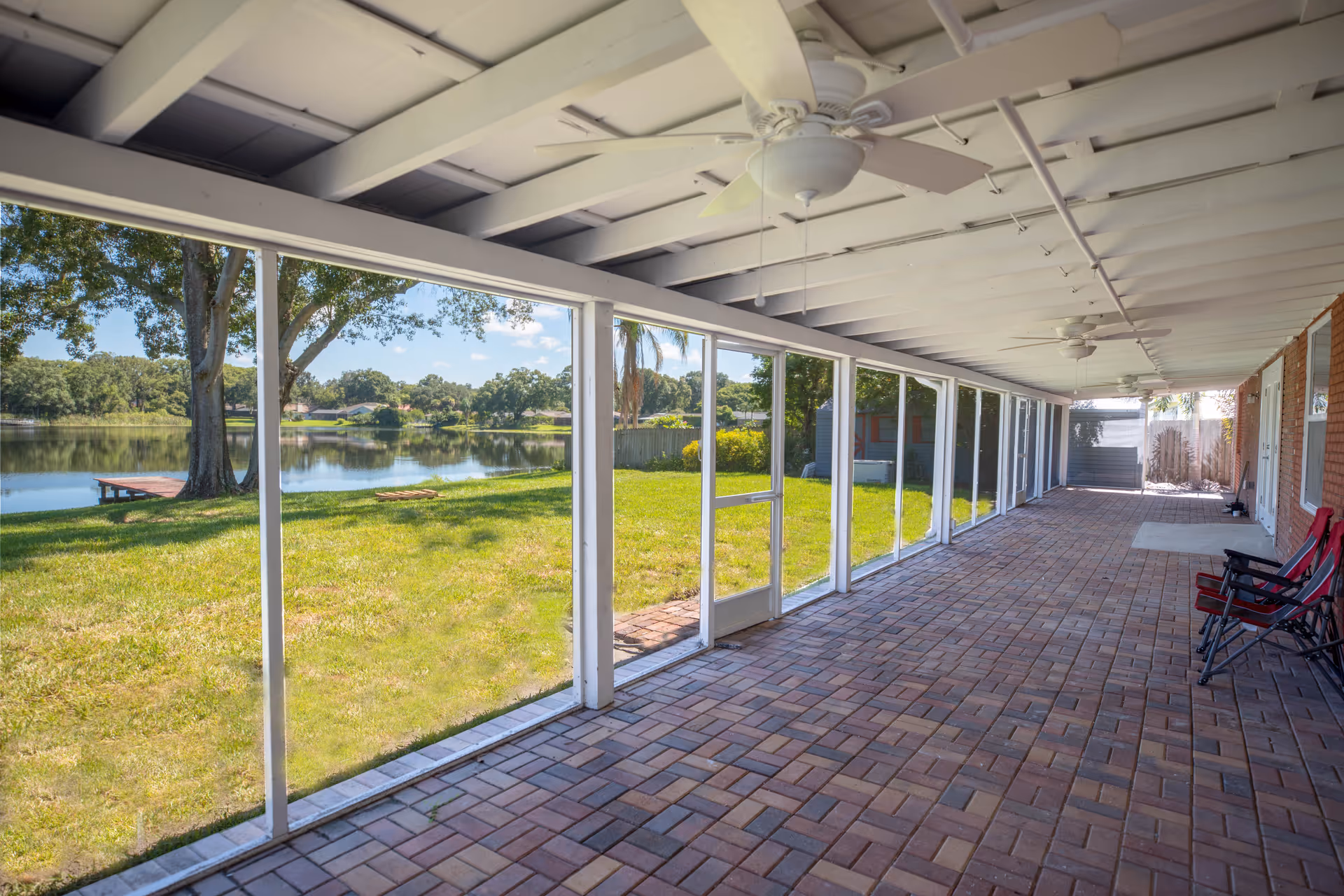 A spacious screened-in porch with brick flooring and ceiling fans, overlooking a grassy yard and a lake with trees and a small dock in the background. There are two red folding chairs placed against the brick wall on the right side.