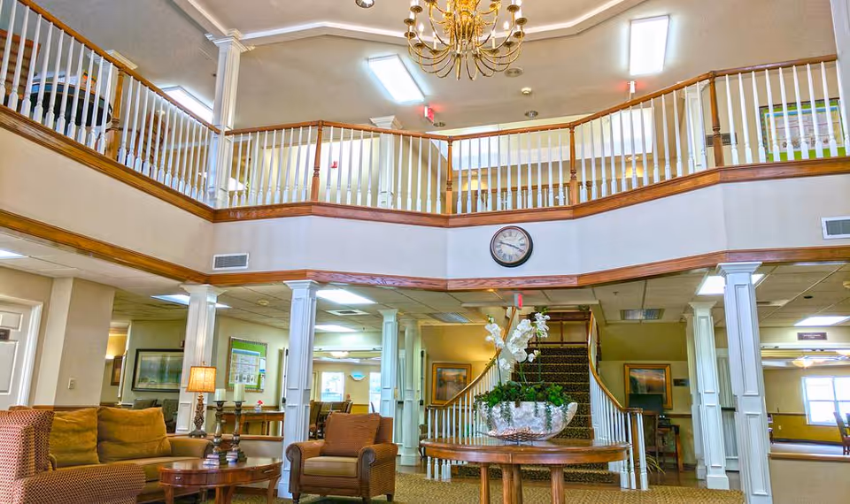 Spacious two-story senior living lobby with seating, central table of flowers, staircase and upper balcony.
