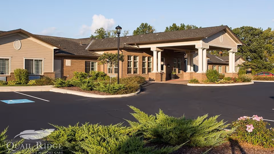 Front entrance of a single-story brick assisted living facility with a covered portico, columns, parking lot, and landscaped beds.