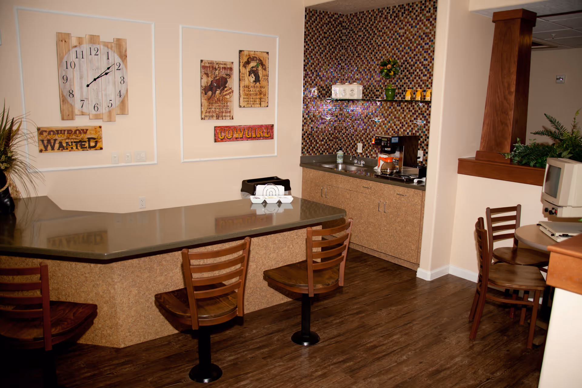 Interior view of a small kitchenette area with a counter and three wooden swivel chairs. The kitchenette has a mosaic tile backsplash, a coffee maker, and a toaster on the counter. On the wall above the counter, there are decorative signs including a large wooden clock and signs that say 'COWBOY WANTED' and 'COWGIRL'. To the right, there is a small round table with two wooden chairs and an old computer on it. The floor is wooden, and there are some plants visible in the corner.