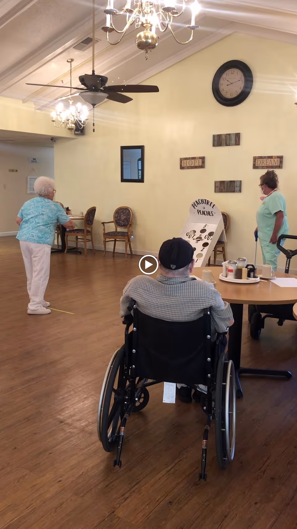 Three elderly individuals in a senior living facility common area. One person is seated in a wheelchair facing a game board labeled 'Peachtree Peaches' while two others stand nearby, one preparing to throw an object. The room has wooden flooring, a ceiling fan, chandeliers, and wall decorations with words like 'Hope', 'Faith', and 'Dream'.