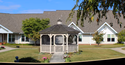 A wooden gazebo sits on a grassy lawn in front of a light-colored building.