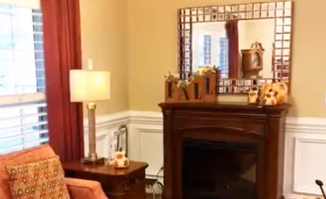 A cozy living room corner featuring a wooden fireplace with decorative items on the mantel, including a clock and autumn-themed decorations. To the left, there is a side table with a lamp and a patterned armchair next to a window with red curtains.