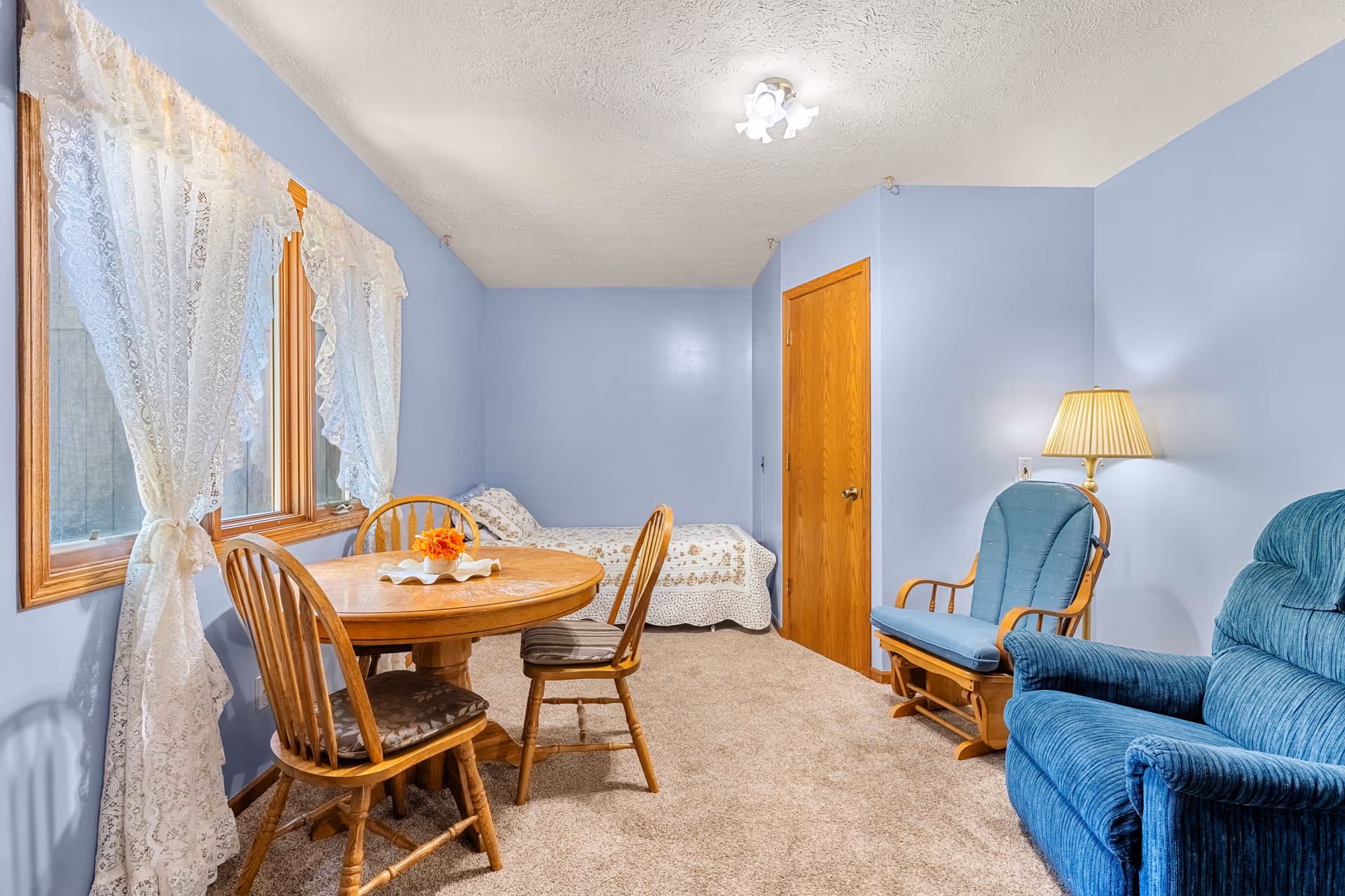 Cozy multipurpose room with blue walls, a round wooden dining table and chairs, a small bed in the corner, and upholstered recliners.