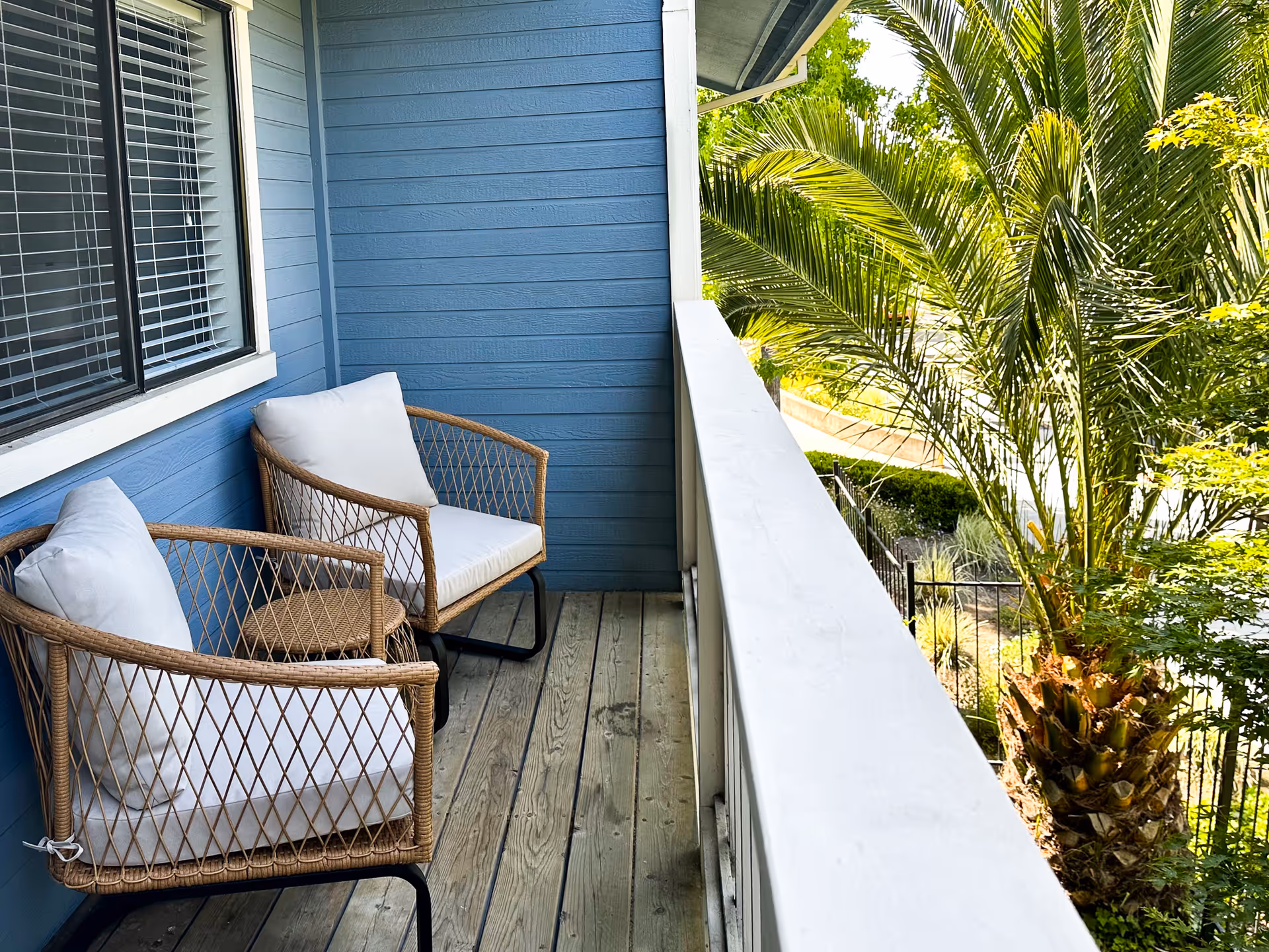 Small outdoor balcony with two wicker chairs with white cushions and a small wicker table between them. The balcony has wooden flooring and blue siding walls. There is a white railing on the right side and green palm trees and other foliage visible beyond the railing.
