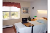 Single-occupancy bedroom with a bed, an overbed table holding a laptop, bedside lamp and phone, a window with a valance, and framed artwork on the wall.