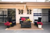 Outdoor patio area with black wicker furniture including a loveseat, two chairs, and a coffee table. The loveseat has colorful cushions and a planter with yellow flowers on the table. There are potted plants with pink flowers on either side of the seating area, and two wall-mounted planters with green plants on the beige wall behind. Sliding glass doors with vertical blinds are visible on both sides of the patio.