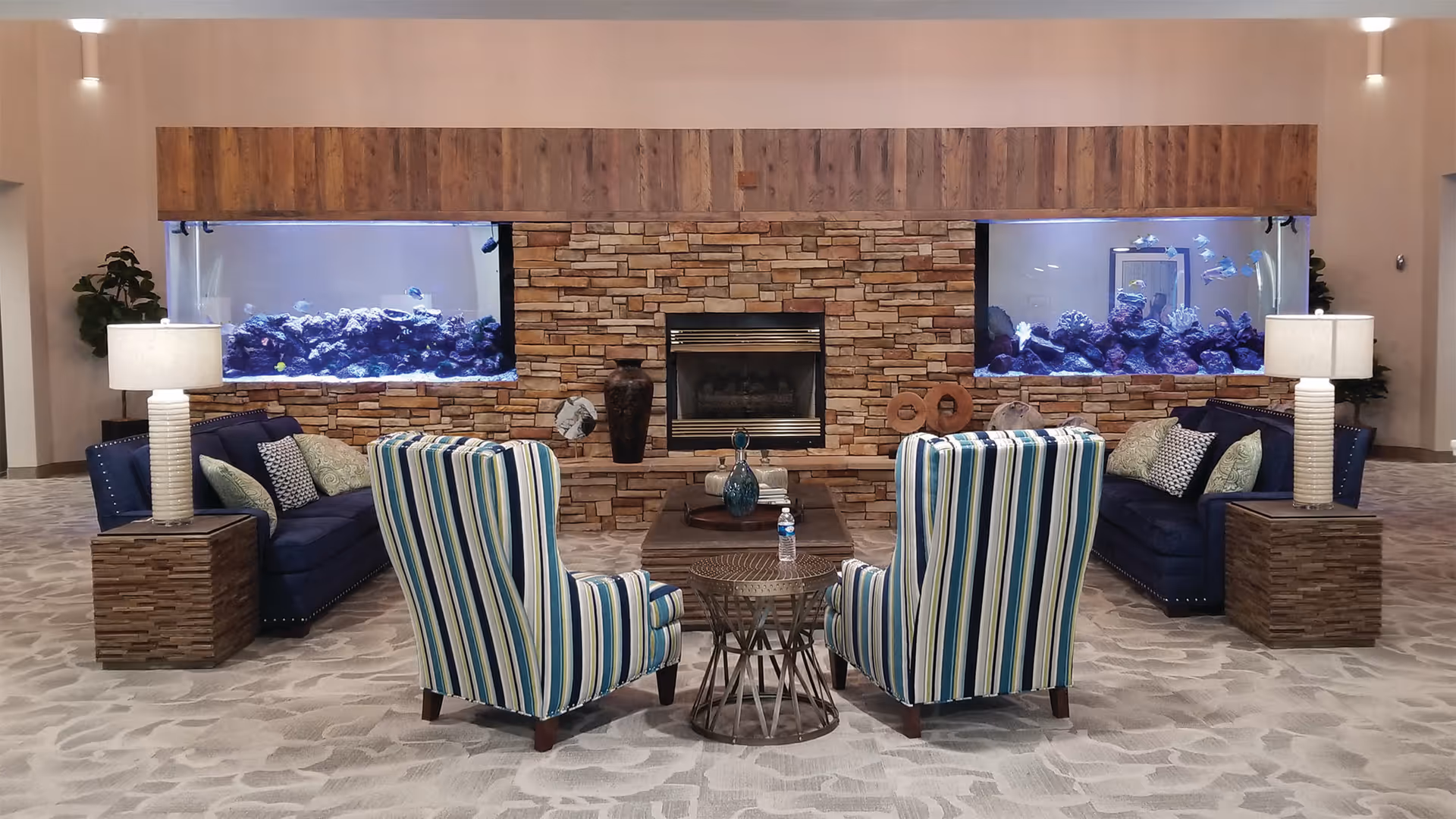 Lobby-style lounge with striped armchairs and sofas facing a stone fireplace flanked by large aquariums.