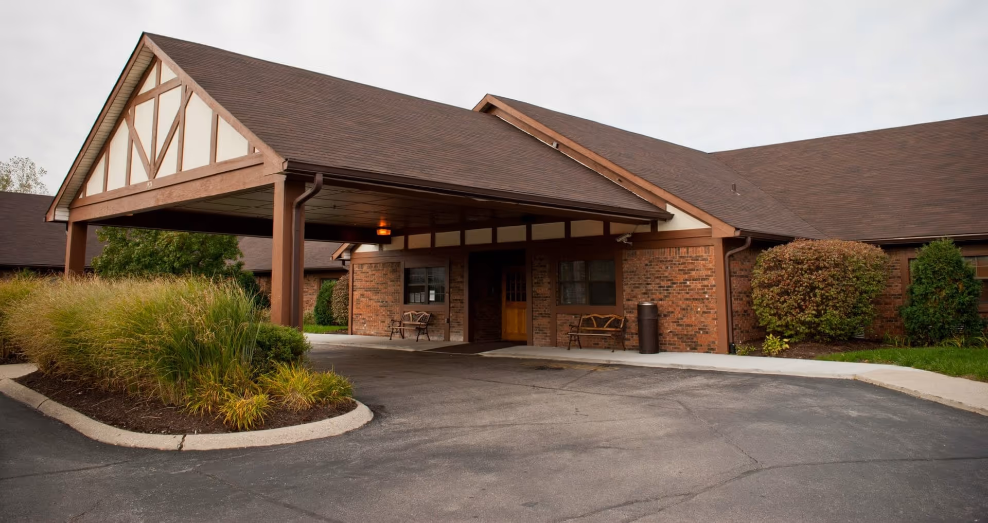 Exterior view of a single-story brick building with a large covered entrance supported by wooden beams. There are two benches near the entrance and landscaped bushes and grass around the driveway.