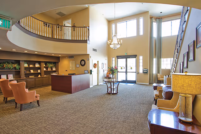 Spacious senior living facility lobby with high ceilings, large windows, and a glass entrance door. The area features a wooden reception desk, several upholstered chairs, a round table with a floral arrangement, and a staircase leading to an upper level. Warm lighting and neutral tones create a welcoming atmosphere.