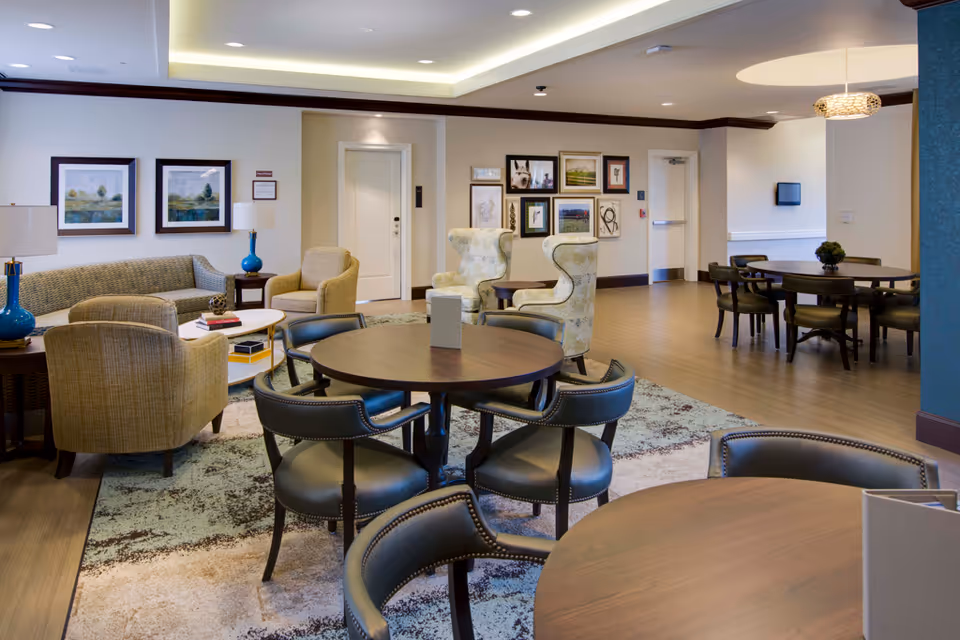 A spacious and well-lit common area in a senior living facility featuring multiple round wooden tables with black cushioned chairs, a seating area with a beige sofa, armchairs, and two patterned wingback chairs. The walls are decorated with framed artwork, and the floor is a combination of wood and area rugs. There are lamps on side tables and a modern light fixture hanging from the ceiling.
