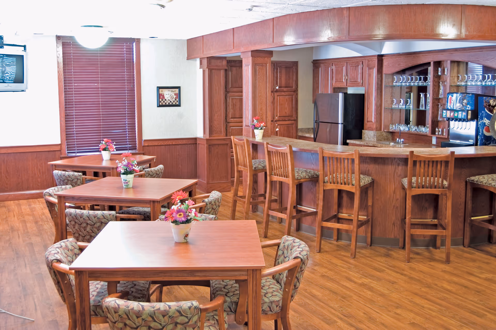 A cozy dining area in Sweetwater Retirement Community featuring wooden tables with floral centerpieces, cushioned chairs with patterned upholstery, a wooden bar with high stools, a refrigerator, and a beverage dispenser. The room has wooden flooring and wood-paneled walls with a window covered by blinds.