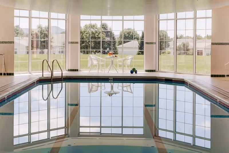 Indoor swimming pool with large floor-to-ceiling windows showing a green outdoor area with trees and houses. There is a small round table with two white chairs and some fruit and dumbbells on the floor near the windows.