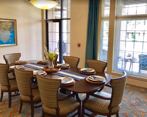 A dining room with a dark wooden table set for eight people. Each place setting includes a plate, napkin, and silverware. The chairs have striped upholstery. A vase with flowers is placed in the center of the table. Large windows with white blinds and teal curtains allow natural light into the room. A glass door and a framed painting are visible on the left wall.